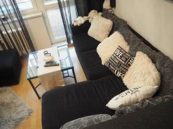 Světlý obývací pokoj s parketovou podlahou, velká okna zajišťují dostatek přirozeného světla, moderní pohovka v šedém odstínu.