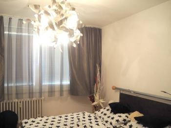 Útulná ložnice s moderním lustrm, velká okna s závěsy, bílými zdmi a jednoduchým dekorem, ideální pro klidný odpočinek.