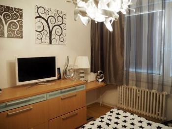 Kompaktní ložnice s parketovou podlahou, velkým oknem a moderním nábytkem. Stylově dekorováno, ideální pro městské bydlení.